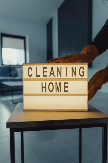 A person wearing orange gloves is placing a wooden block lightbox with the words 'CLEANING HOME' illuminated in black letters on a modern wooden table. The background shows a clean, well-lit room with