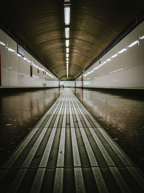 Inside Hampton Wick station, the modern interior features a spacious platform with a high, curved ceiling supported by metal beams and glass panels allowing natural light. The area showcases clean, po