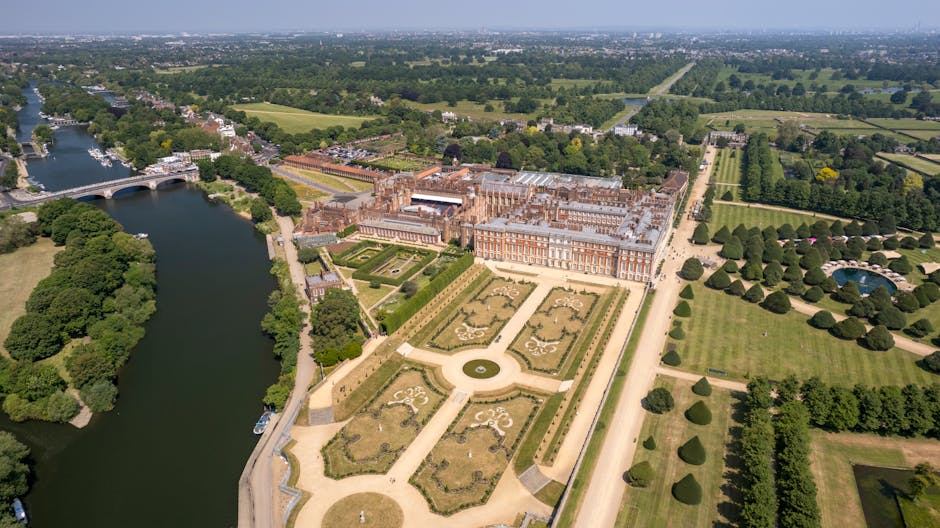 An aerial view of Hampton Court Palace surrounded by its formal gardens and extensive parkland, featuring neatly trimmed hedges, flowerbeds, and tree-lined walkways. The palace's historic red-brick fa