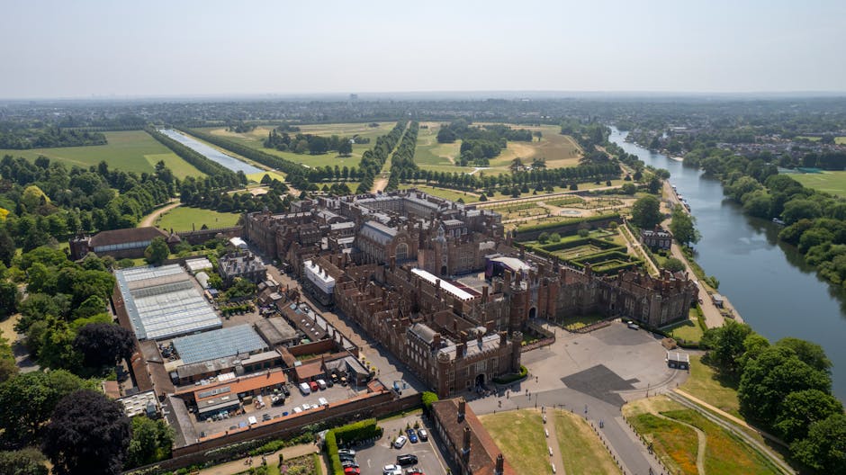 An aerial view of Hampton Court Palace surrounded by its formal gardens and extensive parkland, featuring neatly trimmed hedges, flowerbeds, and tree-lined walkways. The palace's historic red-brick fa